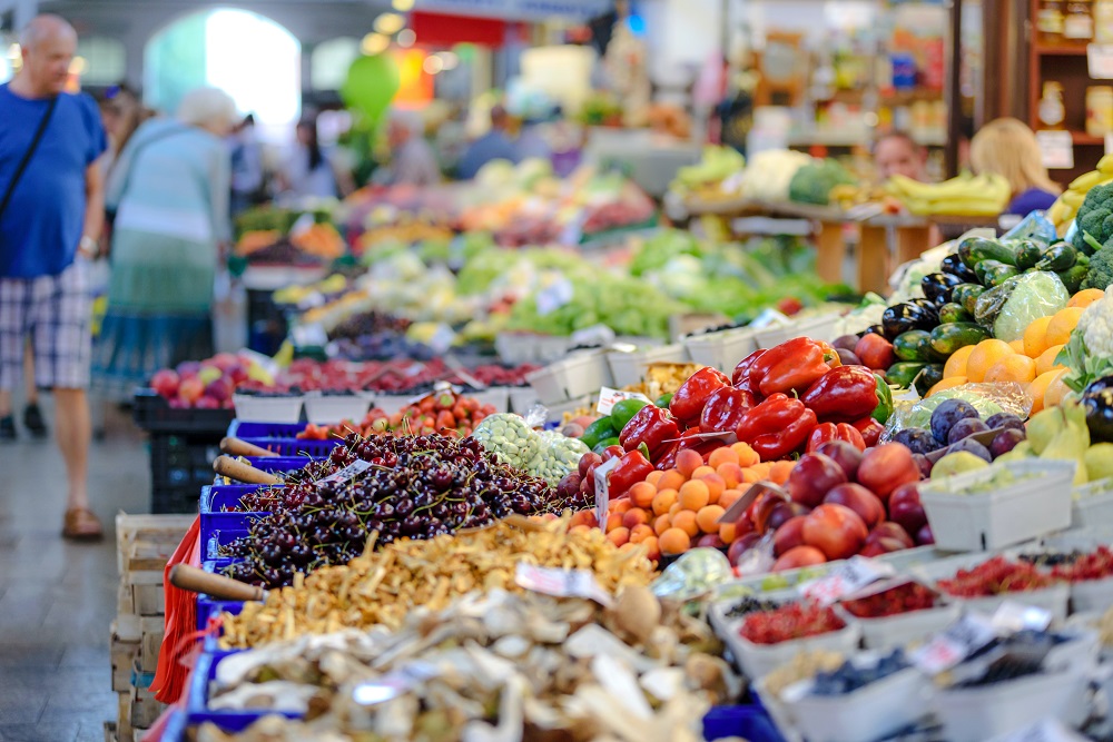 Photo d'un étal de marché avec des fruits et légumes, on aperçoit des passants au fond. Photo de PhotoMIX Company via Pexels.