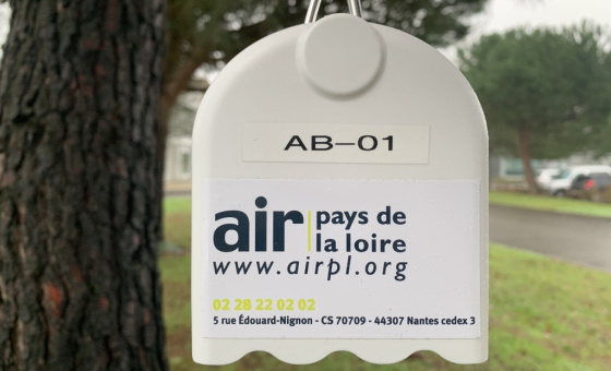 Le capteur à particules fines est un boitier blanc avec un autocollant air pays de la loire. Il est suspendu dehors, devant un arbre