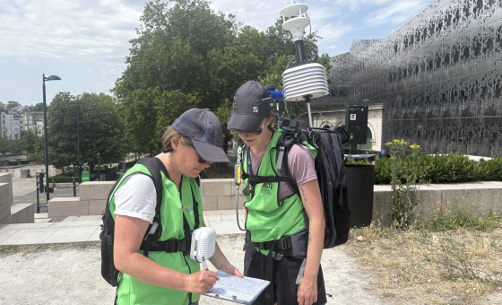 Deux techniciennes portant des gilets verts réalisent une campagne de mesures environnementales en extérieur. L’une prend des notes sur un formulaire tandis que l’autre consulte les relevés d’un équipement de mesure météorologique portable porté sur le dos. La scène se déroule par temps ensoleillé, devant un bâtiment moderne et des arbres.