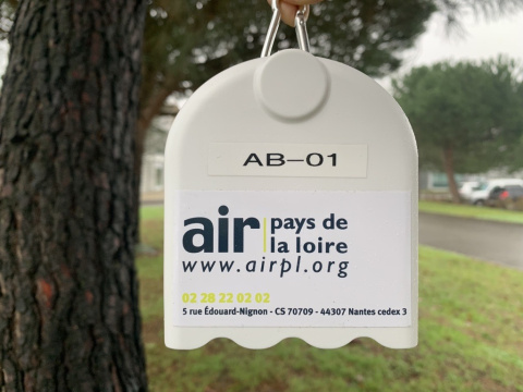 Le capteur à particules fines est un boitier blanc avec un autocollant air pays de la loire. Il est suspendu dehors, devant un arbre