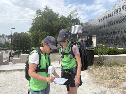 Deux techniciennes portant des gilets verts réalisent une campagne de mesures environnementales en extérieur. L’une prend des notes sur un formulaire tandis que l’autre consulte les relevés d’un équipement de mesure météorologique portable porté sur le dos. La scène se déroule par temps ensoleillé, devant un bâtiment moderne et des arbres.